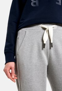 Graue Jogginghose aus weichem Stoff mit weißer Kordel, Seitentaschen und lockerem Schnitt. Kombiniert mit einem marineblauen Sweatshirt.