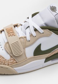 Sneaker featuring white leather, beige accents, and a green Nike swoosh. Textured panel and adjustable strap with embossed branding.