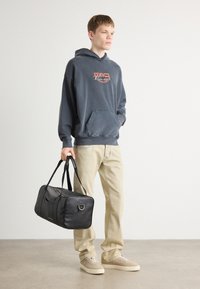 Bolsa de viaje de cuero negra con cremallera superior, asas cortas y un acento metálico. Modelo vistiendo una sudadera azul marino desgastada y pantalones beige claros.