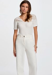 Hvid top med blonde skulderdetaljer, korte ærmer og samlet front. Kombineret med høj talje hvide jeans. Glat tekstur.