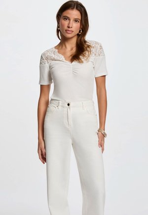 Top bianco con dettagli in pizzo sulle spalle, maniche corte e frontale arricciato. Abbinato a jeans bianchi a vita alta. Texture liscia.