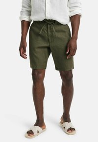Olivgrüne Leinen-Shorts mit einem Kordelzug und Seitentaschen. Kombiniert mit beigen Rutschsandalen für einen lässigen Look.