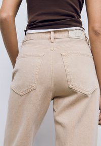 Bež traperice s srednjim strukom, dva stražnja džepa s vidljivim šavovima i suptilnom teksturiranom završnom obradom. Pojas uključuje brendiranje.