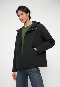 Młody mężczyzna w czarnej kurtce z kapturem na zielonym swetrze i niebieskich dżinsach, pozujący z jedną ręką w kieszeni na jednolitym tle.