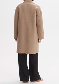 Manteau en laine beige avec un design structuré, coupe légèrement oversize, col cranté et manches longues, associé à un pantalon noir à jambes larges.