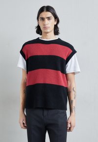 Top sans manches en tricot rayé noir et rouge, présentant une texture côtelée et un col rond, porté sur une chemise blanche.