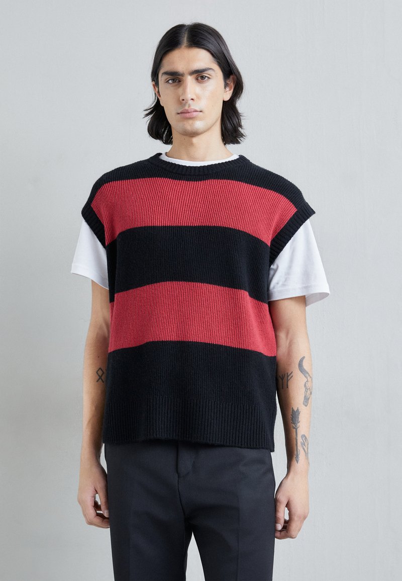 Top sans manches en tricot rayé noir et rouge, présentant une texture côtelée et un col rond, porté sur une chemise blanche.