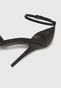 Μαύρο πέδιλο με ψηλό τακούνι που διαθέτει κομψό σχέδιο, λουράκι στον αστράγαλο με αγκράφα, υλικό από λείο δέρμα και μυτερό σχήμα μύτης.