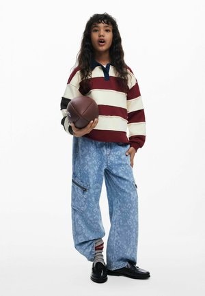 Gestreifte Sweatshirt in Kastanienbraun und Creme mit einem blauen Kragen; blaue gemusterte Cargo-Hosen; hält einen braunen Football; schwarze Schuhe.