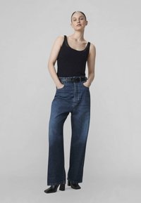 Camiseta sin mangas negra, vaqueros holgados de talle alto, oscuros, con grandes bolsillos y bajos deshilachados, combinados con zapatos de tacón alto negros.