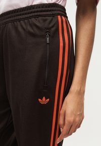 Κοντινό πλάνο ενός ατόμου που φοράει μαύρο αθλητικό παντελόνι με πορτοκαλί ρίγες και λογότυπο Adidas, ελαστική μέση και τσέπη με φερμουάρ στο πλάι.