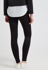 Jeans negros ajustados con cintura alta, hechos de tejido elástico. Presentan bolsillos y una textura suave, combinados con zapatillas blancas.