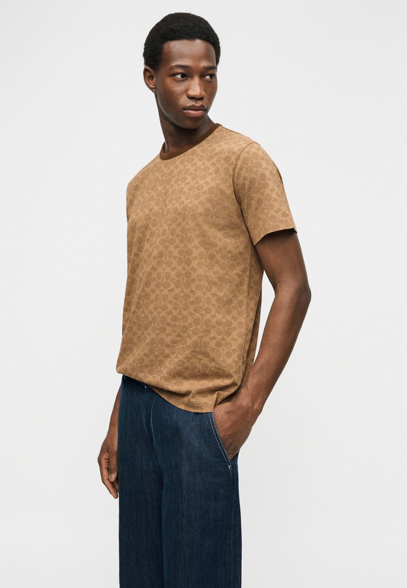 Homme portant une chemise à manches courtes marron à motifs et un jean bleu foncé, debout avec une main dans la poche, regardant sur le côté.