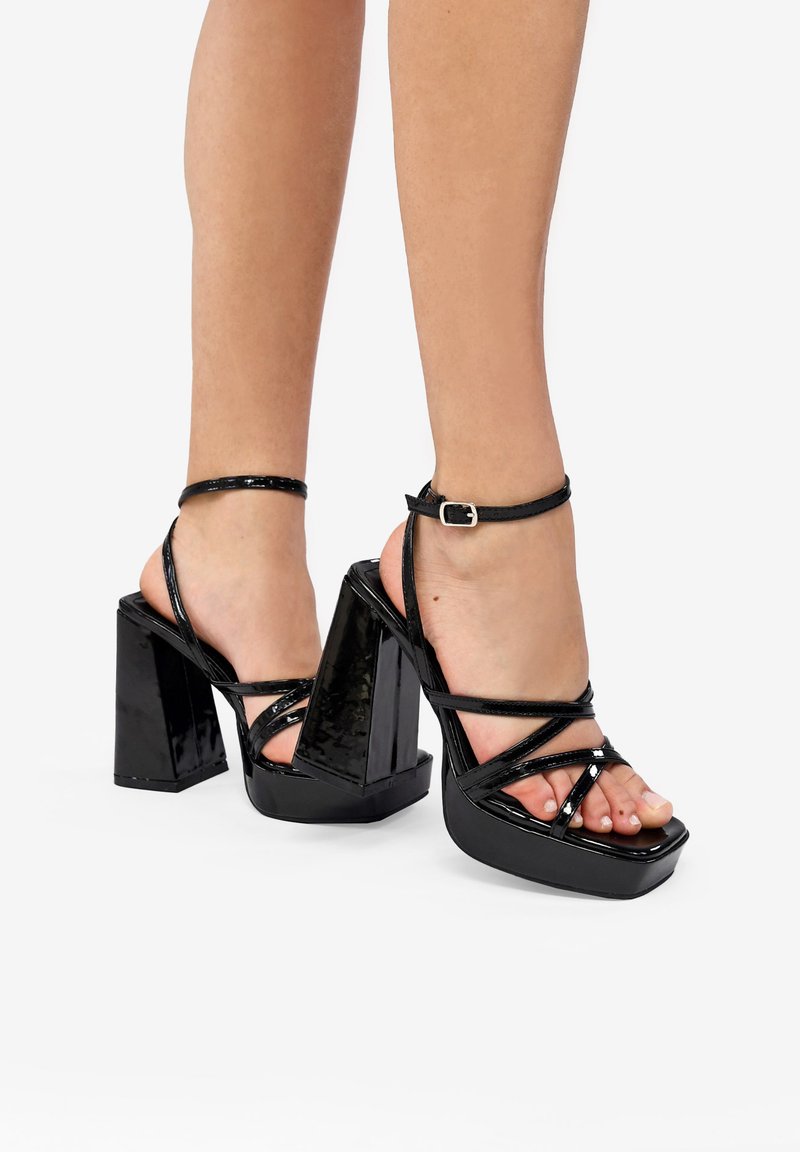 Sandalias de tacón alto de charol negro con un tacón grueso, tira al tobillo y tiras frontales cruzadas, que muestran un diseño de punta abierta.