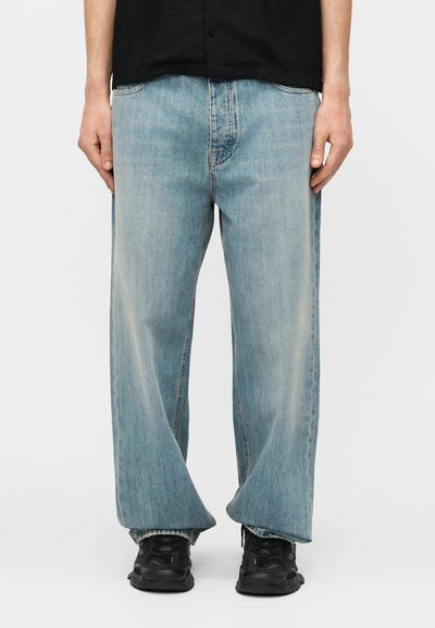 Άνδρας που φοράει ανοιχτό μπλε τζιν με φαρδύ πόδι, μαύρα αθλητικά παπούτσια και μαύρο πουκάμισο με κουμπιά, στέκεται μπροστά σε απλό φόντο.