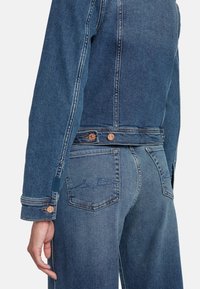Giubbotto in denim di blu medio, con accenti di bottoni in rame e cuciture dettagliate lungo le cuciture e i polsini. La tasca posteriore presenta un design sobrio.