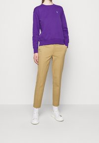 Person med lila långärmad sweatshirt, beige raka byxor, vita strumpor och vita sneakers står med händerna i fickorna.