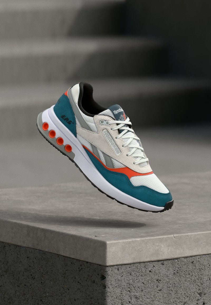 Białe, turkusowe i pomarańczowe sneakersy Reebok z sznurowadłami, teksturowaną podeszwą i markowymi detalami unoszące się nad betonowym występem z zamglonym tłem schodów.
