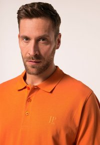 Orange polotröja i bomull med krage, tre knappar och en broderad logotyp på bröstet. Slät textur, avslappnad design.