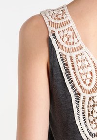 Grå ärmlös topp med en detaljerad krämfärgad spetsdetalj på baksidan med en scalloped mönster och öppen vävdesign.