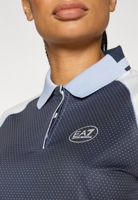 Tumši zils polo krekls ar gaiši ziliem akcentiem, teksturētu audumu un baltām polka punktām. Aprīkots ar pogātu apkakli un logotipu uz krūtīm.