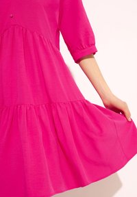 Jasnoróżowa sukienka z krótkimi rękawami i falbaną, wykonana z lekkiego materiału. Na korpusie widoczny jest pojedynczy guzik.
