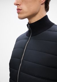 Melnā pufīgā jaka ar augstu ribotu apkakli, kurai ir sudraba rāvējslēdzis un vertikāls quilted raksts. Gluda tekstūra, pielāgota piegriezums.