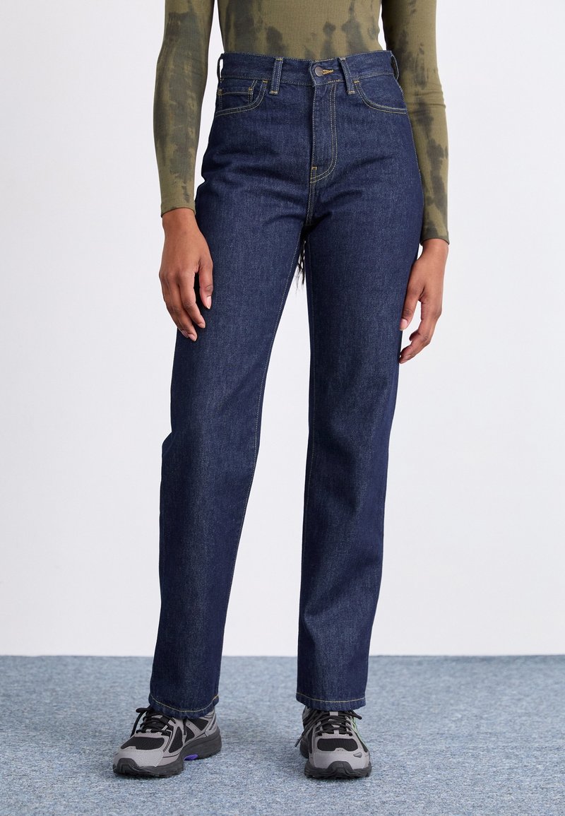 High-waisted dark blue denim jeans with straight leg design, visible stitching, and distressed hem. Paired with gray and black sneakers.