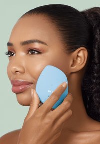 Dispositif de nettoyage facial en silicone bleu tenu contre la joue, avec des boutons à l'avant. Texture lisse, forme ergonomique et design compact.