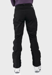 Personne portant un pantalon de ski noir et des bottes de ski blanches vue de dos sur un fond uni.
