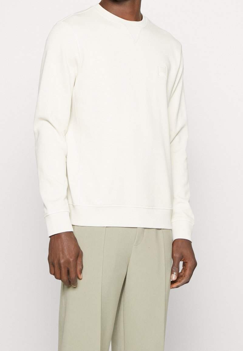 Personne portant un sweat-shirt blanc uni à manches longues avec col rond et un pantalon beige clair, debout devant un fond clair uni.