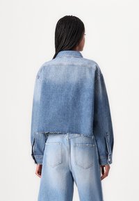 Giacca di jeans tagliata in azzurro chiaro con orlo sfrangiato, polsini con bottoni e colletto classico; abbinata a jeans chiari a vita alta.