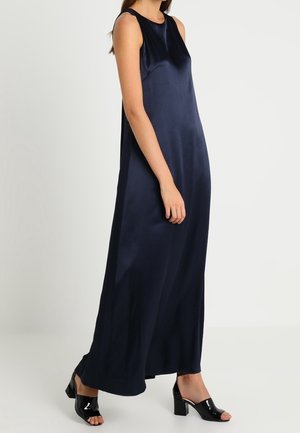 Femme portant une robe longue en satin bleu marine sans manches avec des chaussures noires à talons blocs ouverts à l'avant, debout devant un fond uni.
