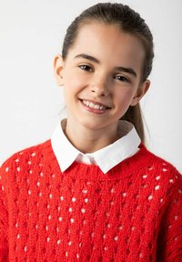 Czerwony sweter dziewiarski z ażurowym wzorem, z białą koszulą z kołnierzykiem pod spodem. Wzór jest teksturowany i przewiewny.