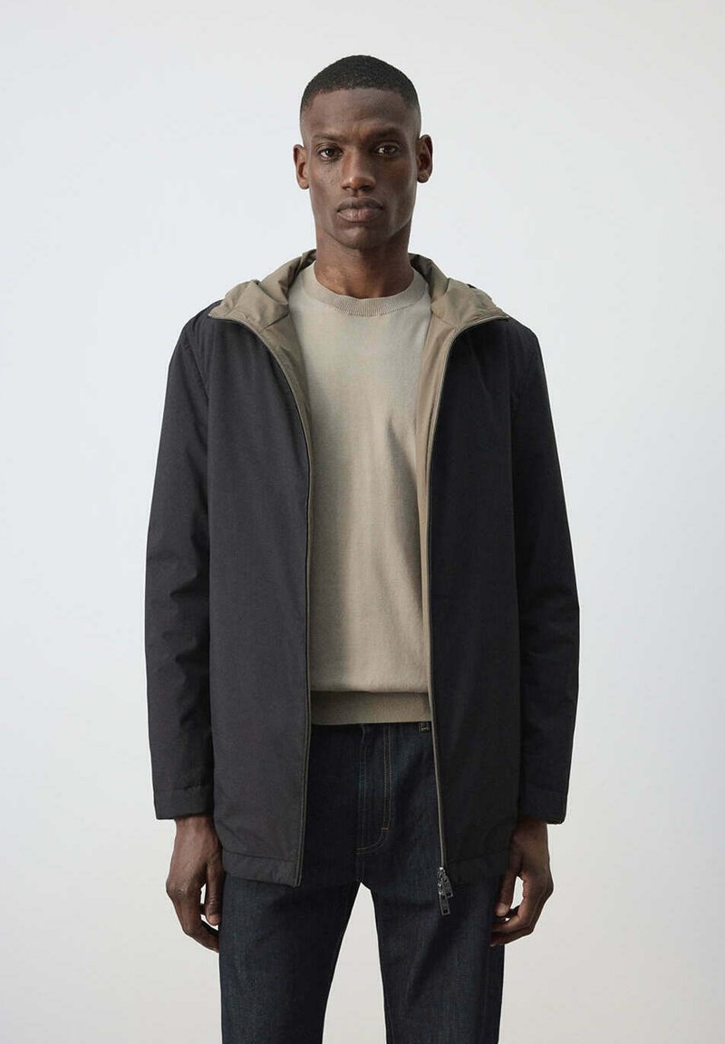 Homme portant une veste noire à fermeture éclair sur un pull beige et un jean foncé, debout devant un fond clair uni.