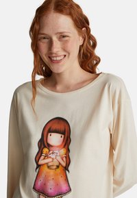 Junge rothaarige Frau lächelt und trägt ein cremefarbenes Shirt mit einer bunten Mädchenillustration, die ein leuchtendes Glas hält.