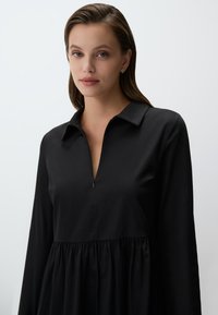 Zwarte lange-mouwen jurk met een kraag en een halve rits. Geverkte taille voegt vorm toe; gladde stof heeft een matte afwerking.