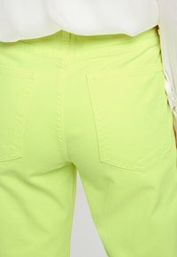 Pantalones de algodón de un brillante color verde lima con bolsillos traseros visibles y tejido texturizado. La cinturilla se encuentra a la altura de la cintura natural.