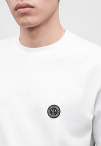 Homme portant un t-shirt blanc avec un petit logo rond noir et blanc Armani Exchange sur la poitrine gauche.