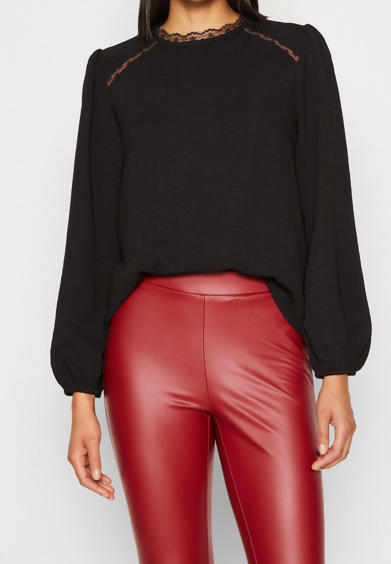 Blouse noire à manches longues avec des détails en dentelle sur les épaules et le col, associée à un pantalon moulant rouge en similicuir. Tissu doux et texturé.