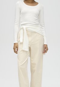 Person wearing a white long-sleeve top with a tied waist detail and loose beige trousers against a plain light background.
