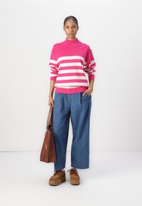 Pull rayé rose et crème avec un col montant et des boutons, associé à un pantalon en denim bleu large et des chaussures à plateforme marron. Tient un sac marron.