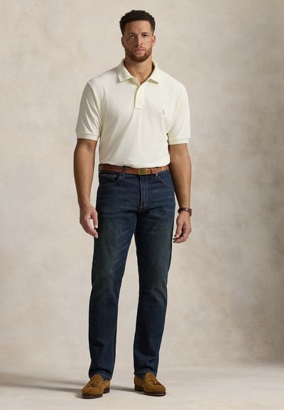 Šviesiai geltonas polo marškinėlis; tamsiai mėlyni džinsai; rudas odinis diržas; rudi zomšiniai loaferiai; stovinti poza; neutrali fone.