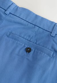 Gros plan sur un tissu bleu montrant la poche arrière d’un pantalon avec un seul bouton noir et des passants de ceinture à la taille.