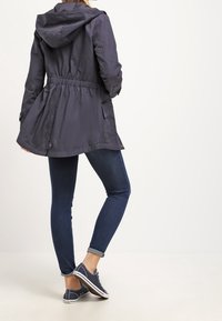 Veste à capuche bleu marine avec taille resserrée, manches longues et poches latérales, associée à un jean foncé et des baskets bleu marine. Texture lisse.