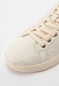Gros plan sur l'avant d'une basket beige avec un motif texturé de peau de serpent et des lacets assortis sur fond blanc.