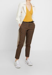 Bruna rutiga högmidjade byxor med ett svart bälte, i kombination med en senapsgul topp och en krämfärgad trenchcoat. Vita sneakers fulländar looken.