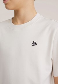 Světlé béžové bavlněné tričko s kulatým výstřihem a krátkými rukávy, s malým černým vyšitým logem ptáka na hrudi.