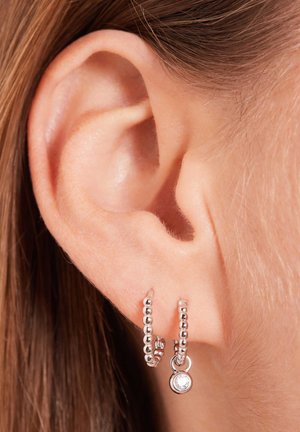 Silver hoop earrings with a beaded design and a small dangling charm featuring a clear gemstone, displayed on an ear close-up.