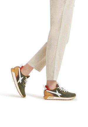 Zapatillas verde y marrón con acentos blancos, talón rojo y suela texturizada. Presentan un diseño de cordones y un logo visible en el lateral.